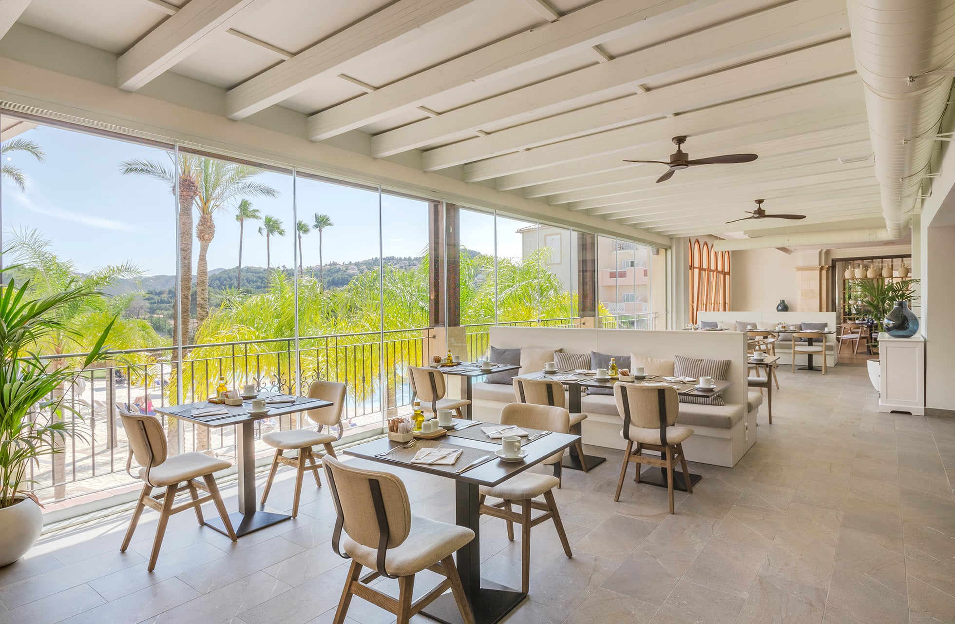 lys og moderne hotellrestaurant med dekkede bord, store vinduer og åpen terrasse mot grøntområder og palmer i dagslys
