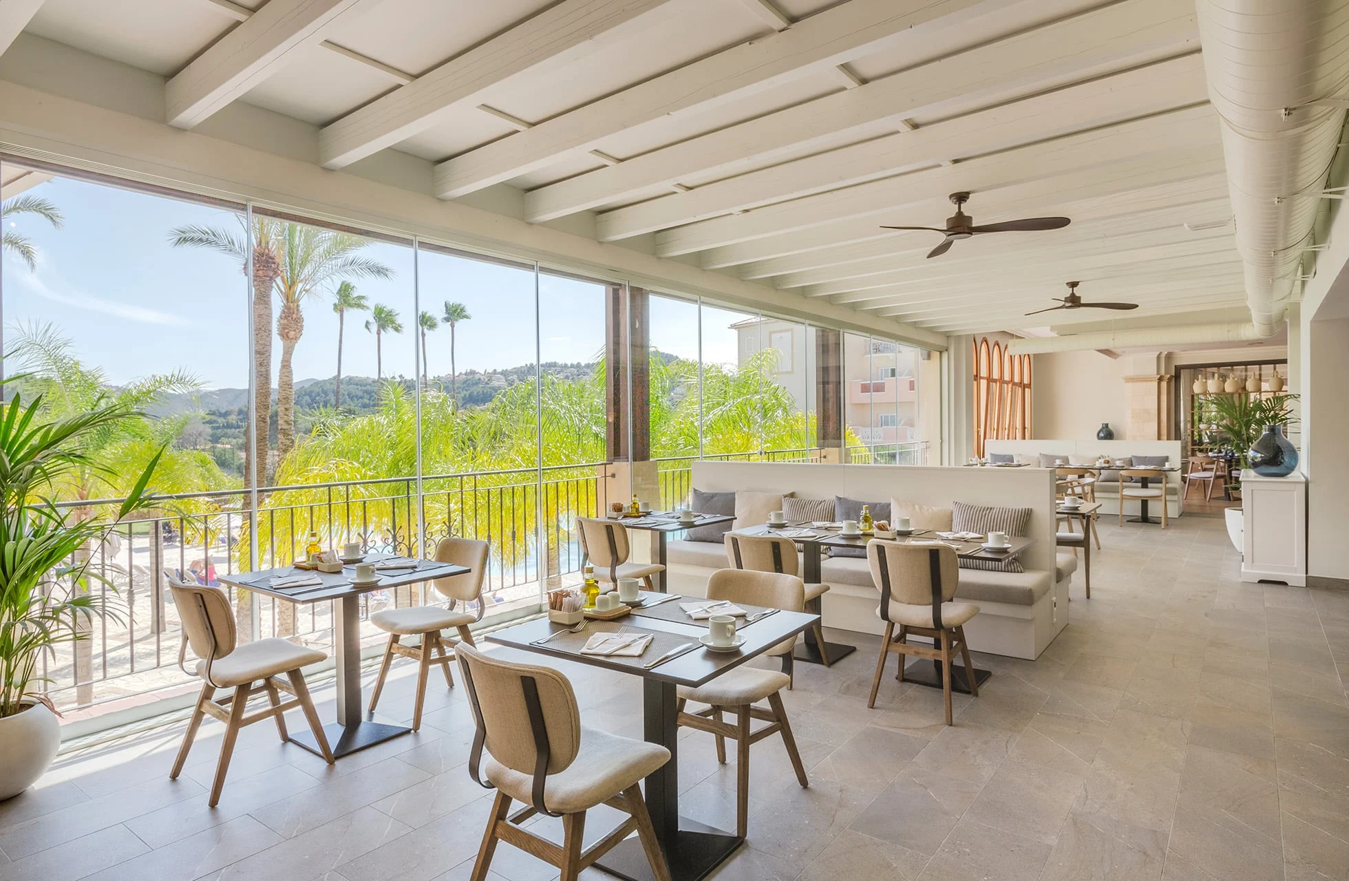 lys og moderne hotellrestaurant med dekkede bord, store vinduer og åpen terrasse mot grøntområder og palmer i dagslys