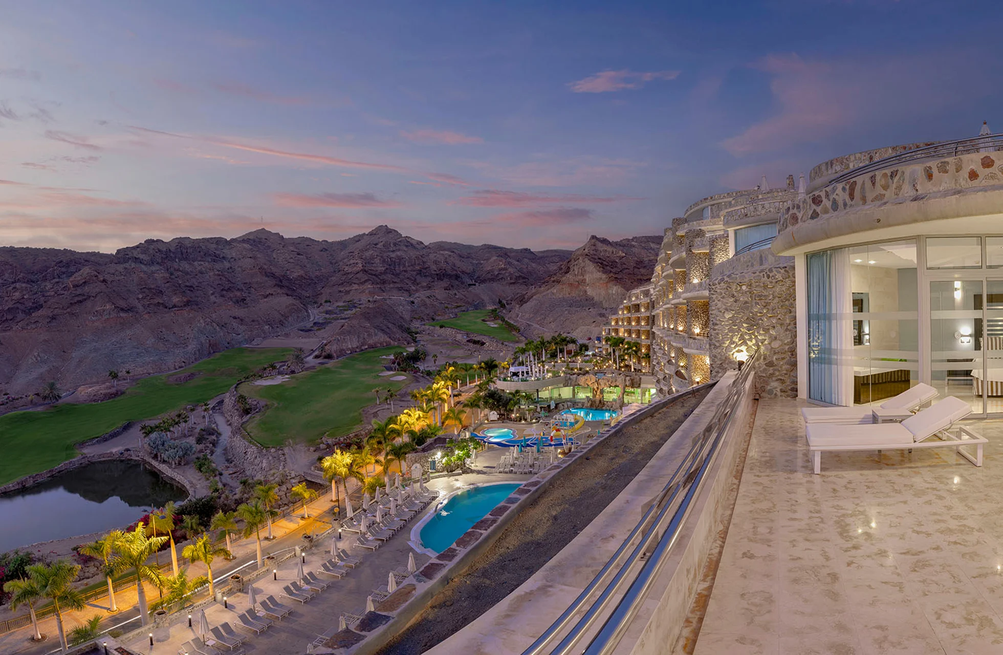 utsikt fra terrasse over anfi emirald club i skumring, med opplyste bassenger, palmer, hotellbygning og kupert landskap med golfbane