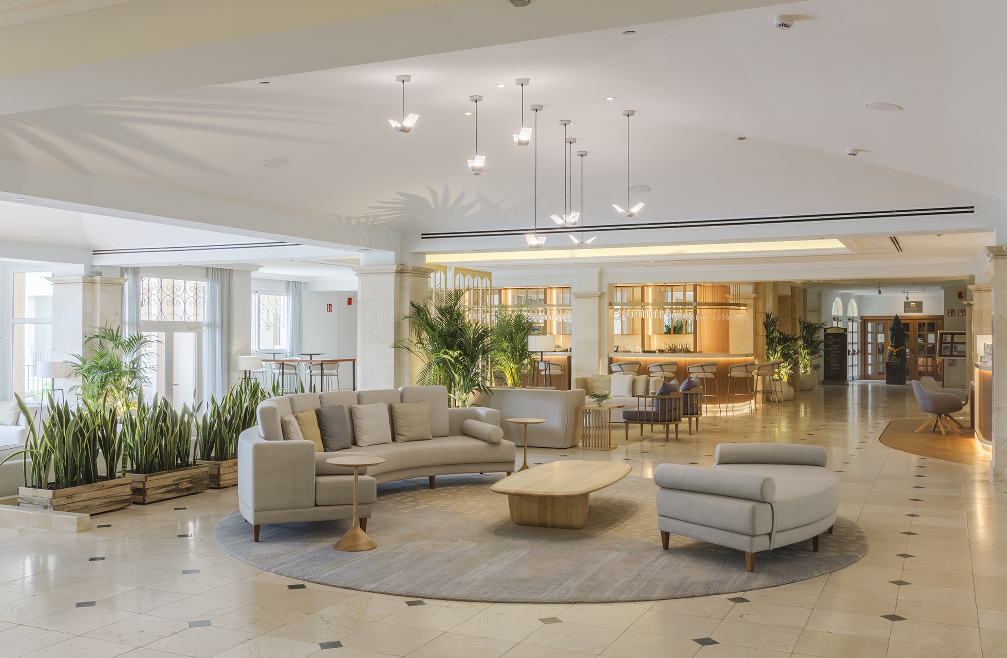 romslig og lys hotellobby med flere sittegrupper, sofaer og lenestoler, store potteplanter, marmorgulv samt resepsjons- og barområde i bakgrunnen i varm belysning
