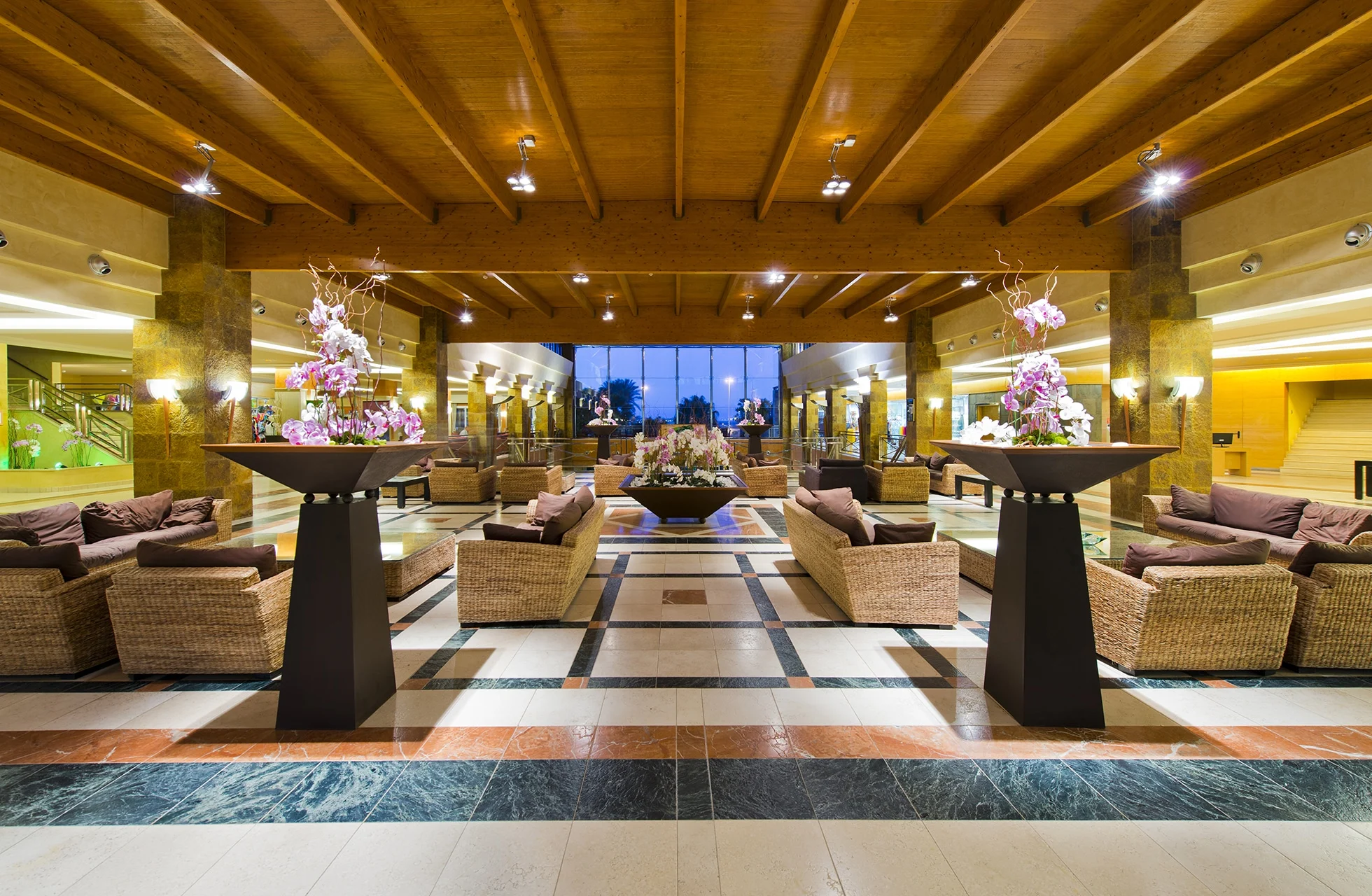 romslig hotellobby med sittegrupper, lenestoler, blomsterarrangementer og tretak i symmetrisk og elegant miljø