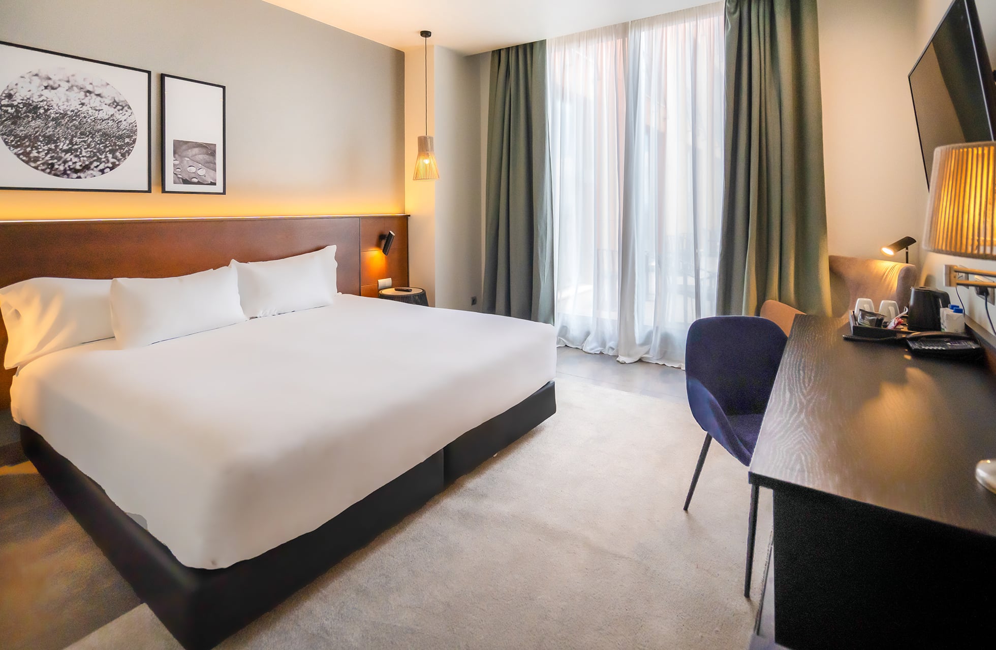 moderne hotellrom med dobbeltseng, nattbord og veggbelysning, skrivebord med stol og tv på veggen, store vinduer med gardiner som slipper inn dagslys