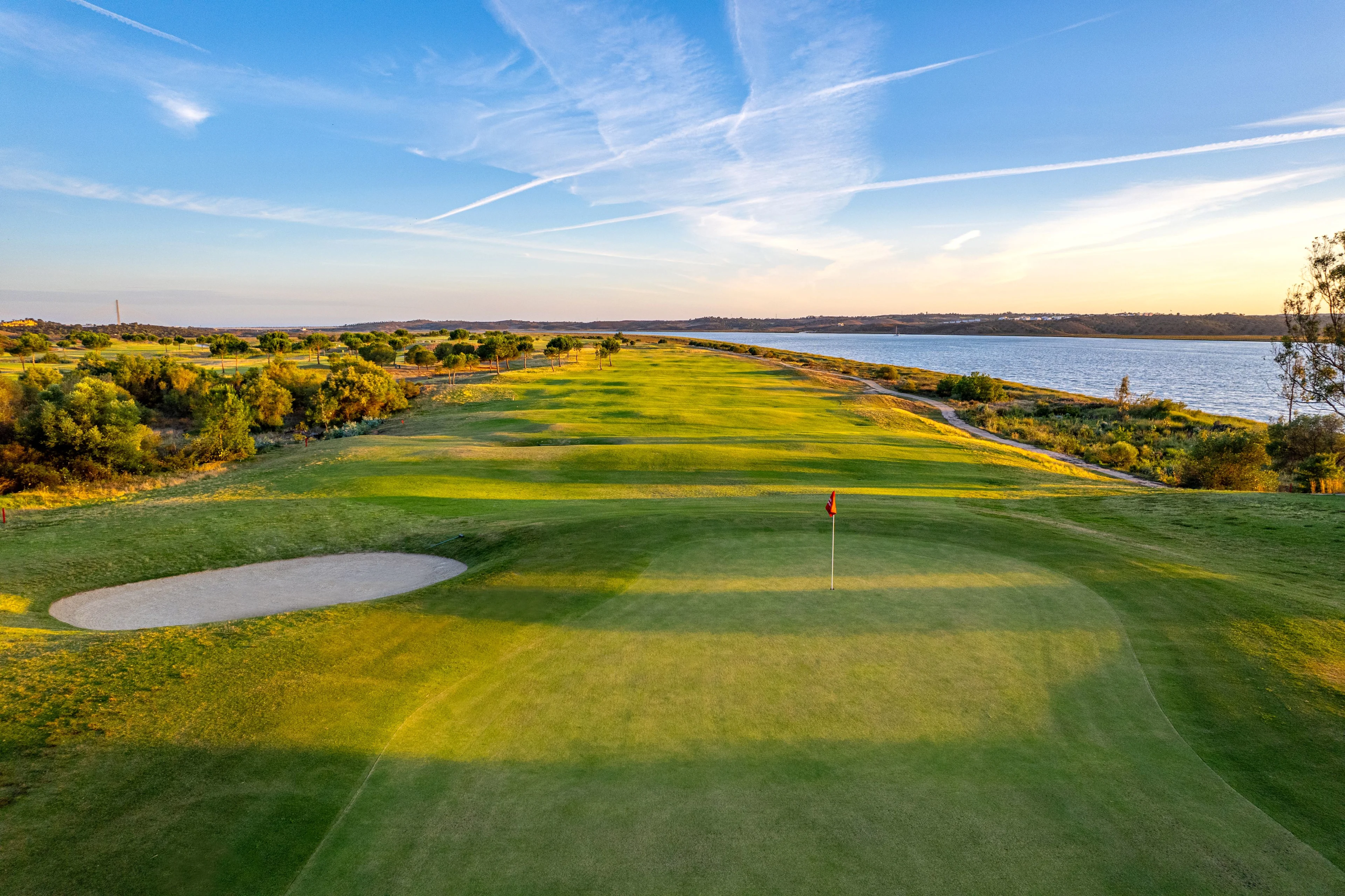 linksinspirerad golfbana med green, flagga och bunker intill vatten och öppet kustlandskap i kvällsljus