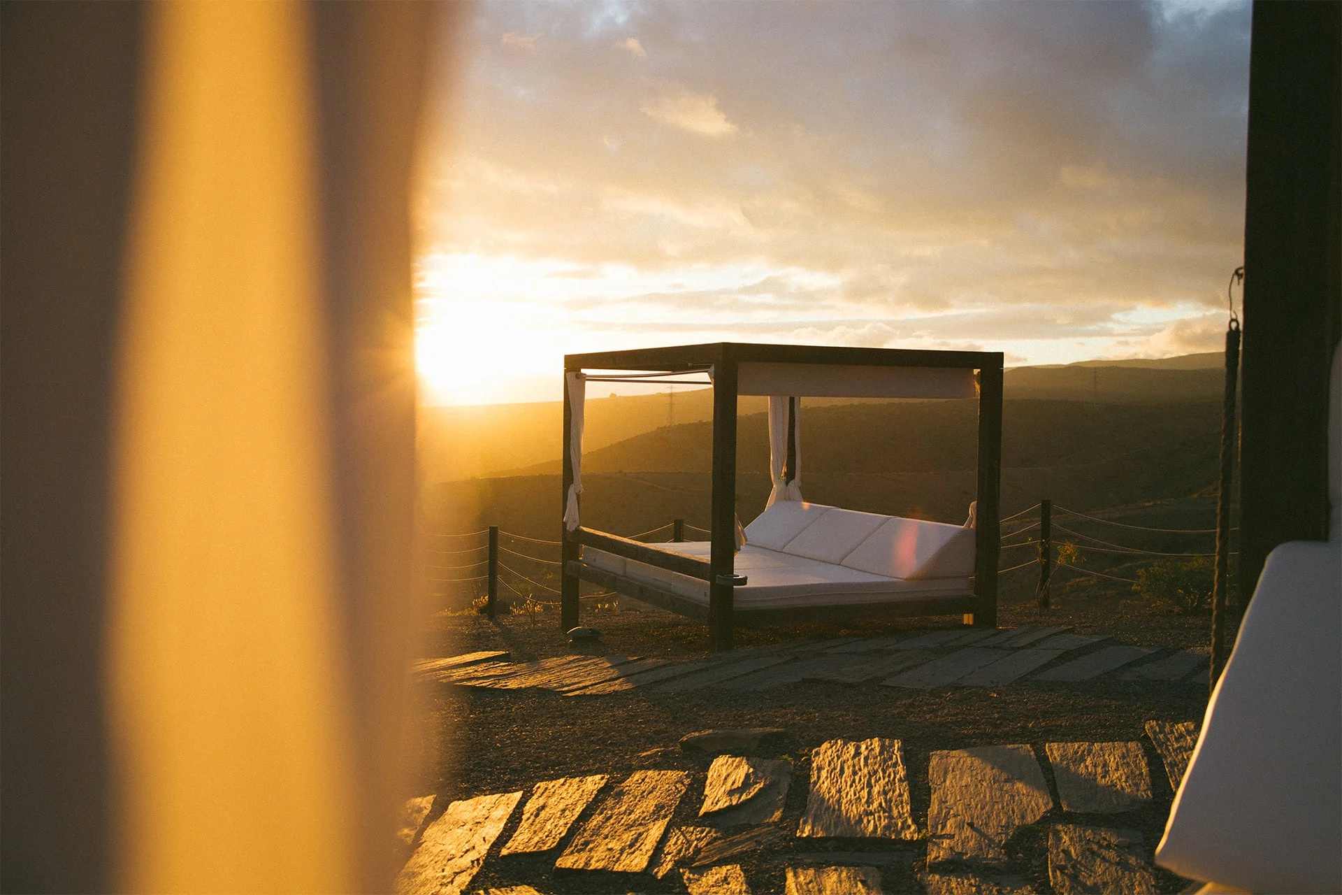 avskjermet dagseng med baldakin på salobre hotel resort i solnedgang, med hvite tekstiler, steinlagt underlag og utsikt over kupert landskap i varmt kveldslys