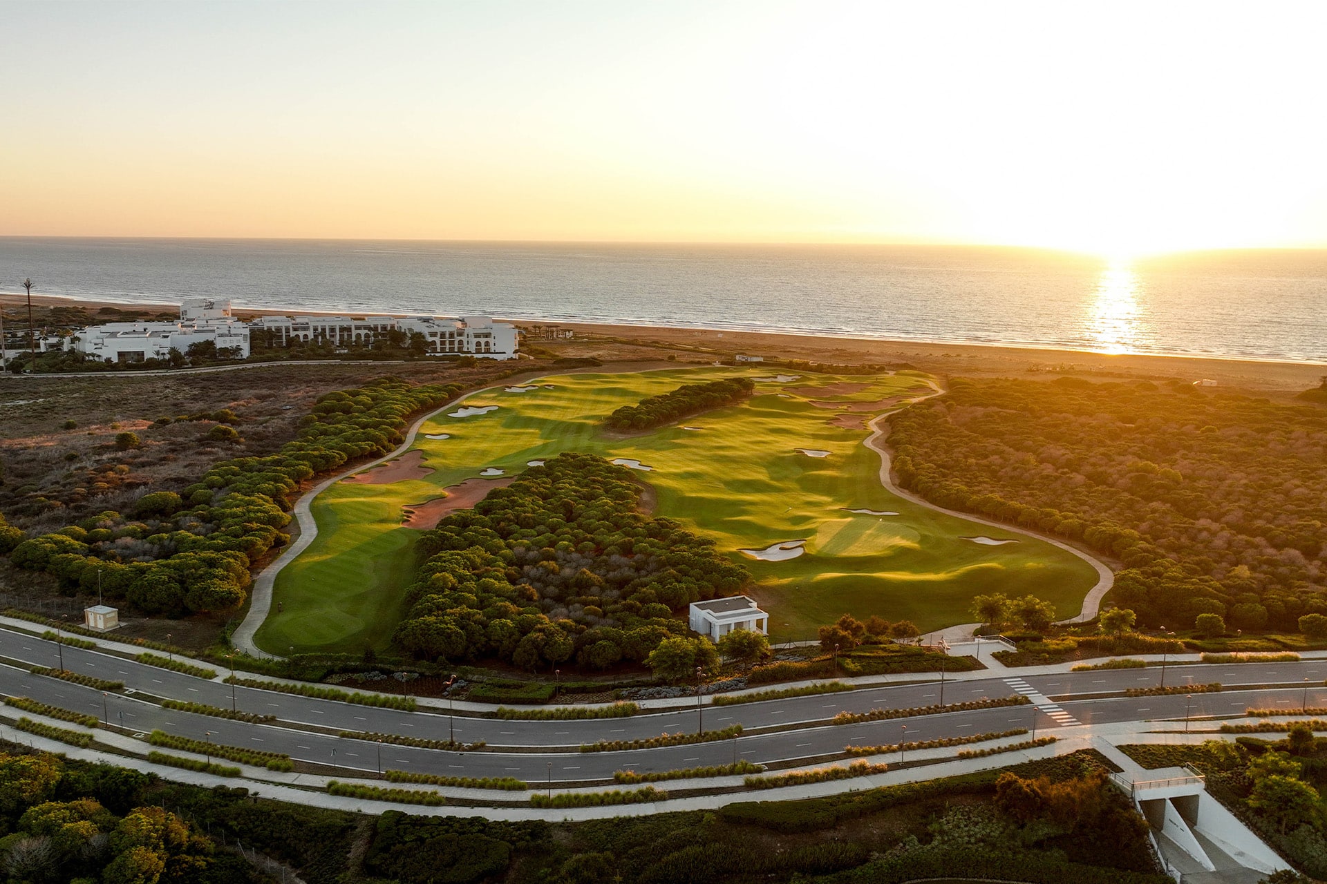 luftfoto af en golfbane med green og fairways omgivet af naturlig vegetation, havet i baggrunden og lav sol ved horisonten i varmt aftenslys