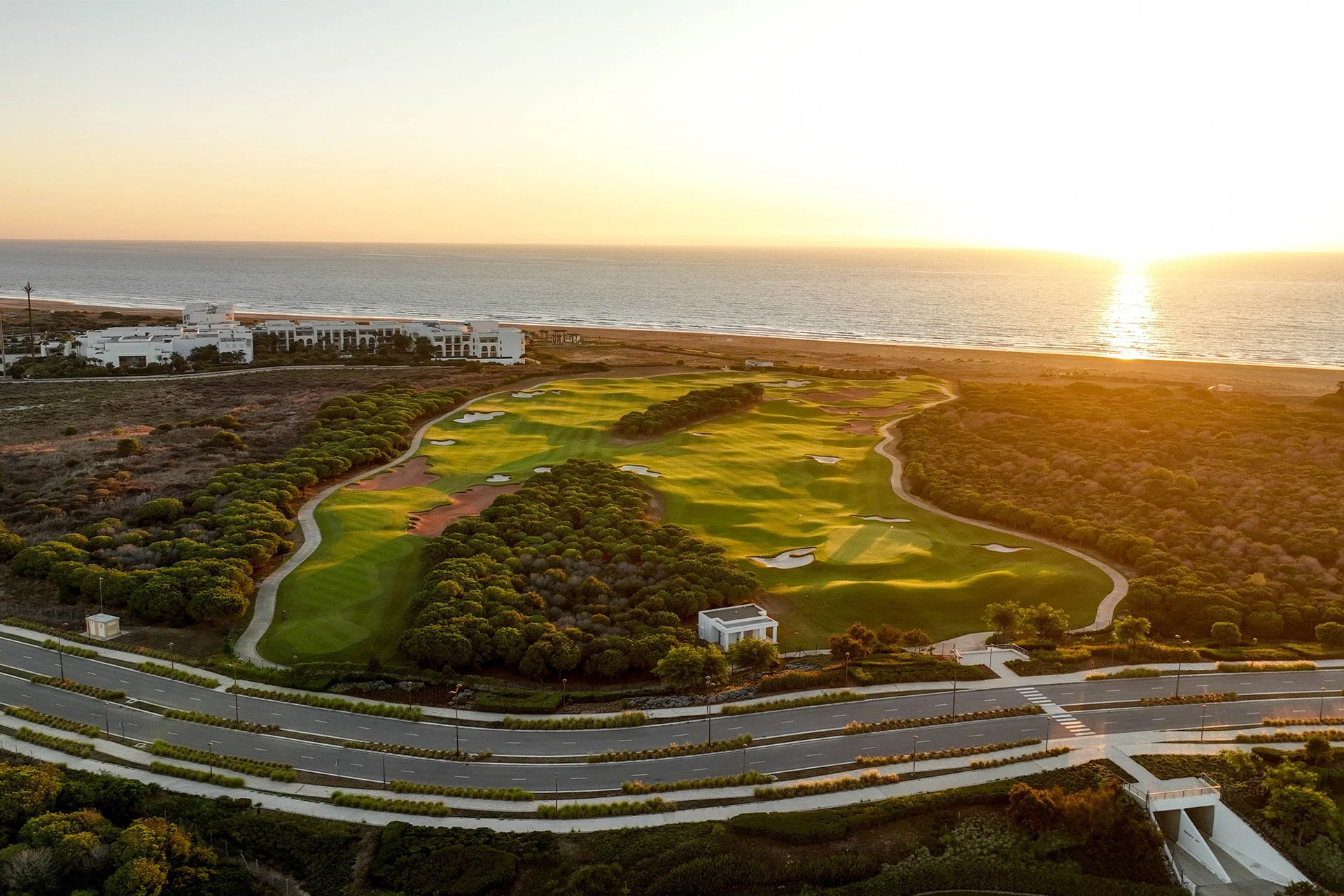 luftfoto af en golfbane med green og fairways omgivet af naturlig vegetation, havet i baggrunden og lav sol ved horisonten i varmt aftenslys