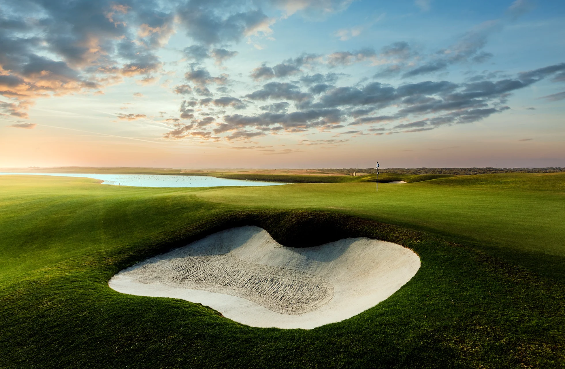 stor hvit sandbunker i forgrunnen omgitt av velstelt fairway, med vannhinder og åpent golflandskap under dramatisk himmel i bakgrunnen