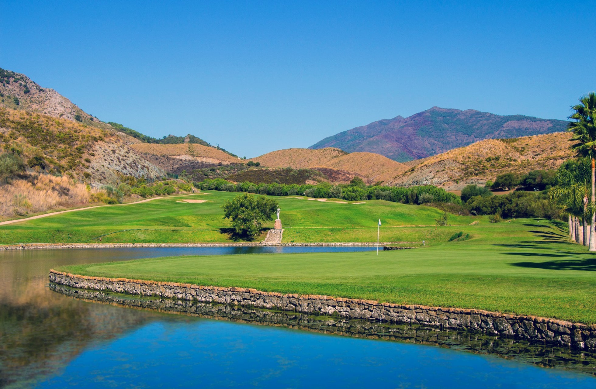golfgreen på villa padierna golf ved vannparti med steinmur i forgrunnen, omgitt av fairway og kupert landskap med fjell i bakgrunnen i klart dagslys