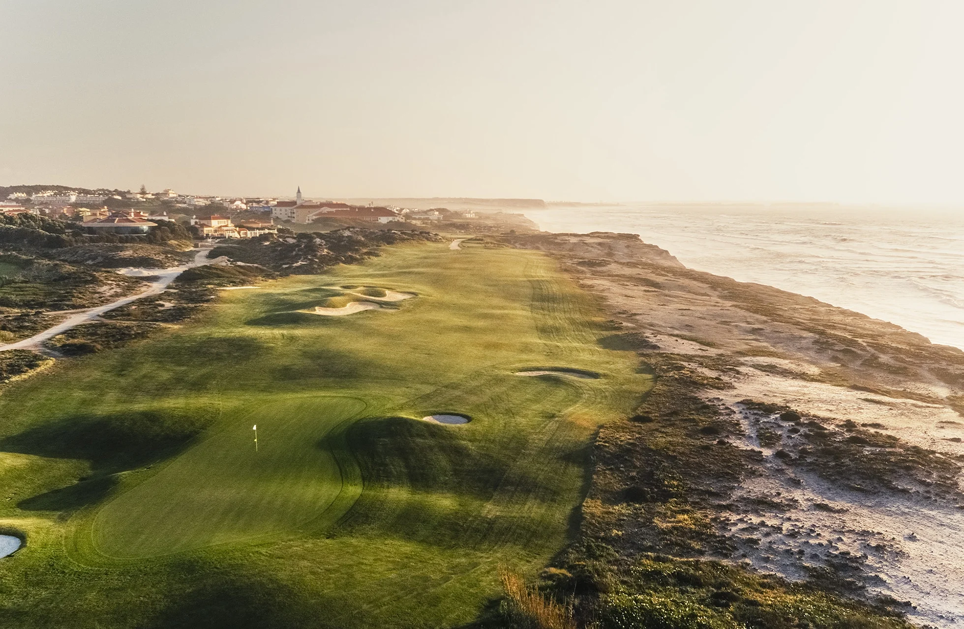vidstrakt golfbane med green, fairway og flere bunkere langs klippekyst, med havet på den ene siden og lav sol som gir lange skygger