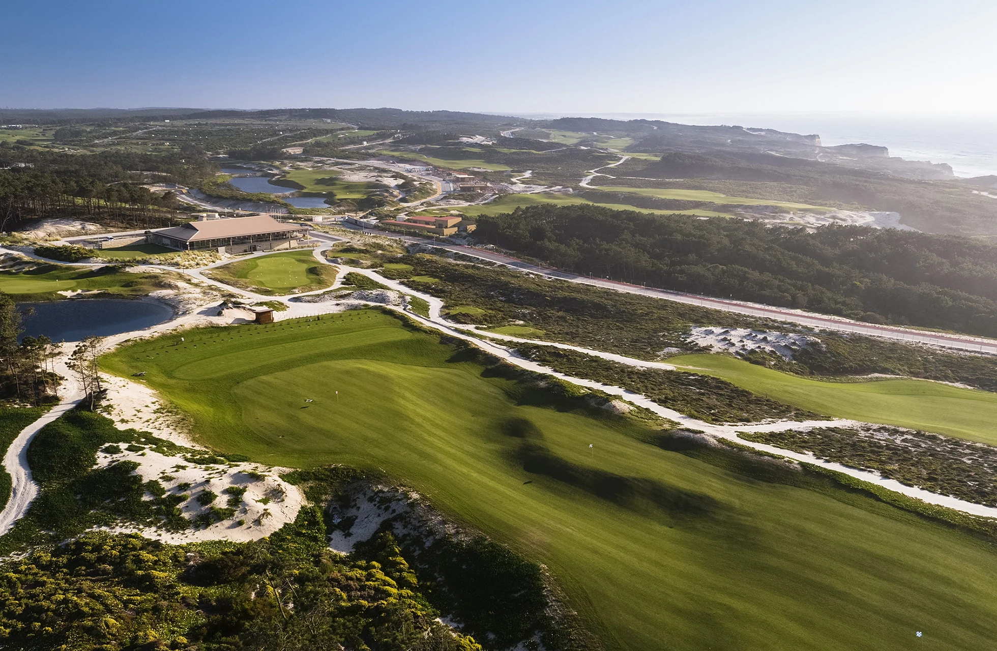 flyfoto av en vidstrakt golfbane med green, fairwayer og bunkere som slynger seg gjennom naturlig vegetasjon, med veier, bygninger og kystlinje synlig i bakgrunnen i klart dagslys
