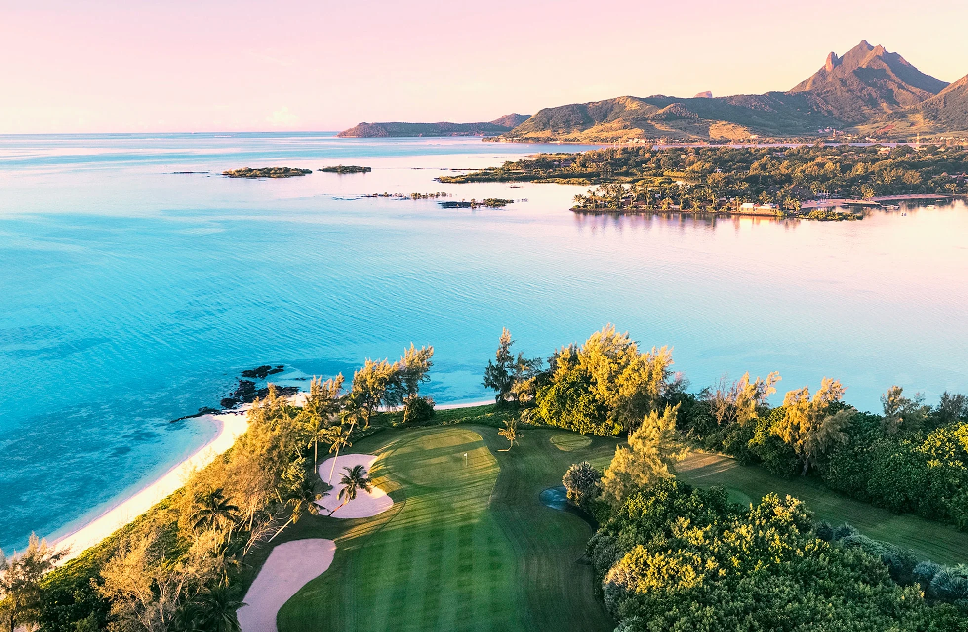 luftfoto af et golfhul på ile aux cerfs golf club med green og fairway omgivet af træer, lagune og hav samt bjerge i baggrunden
