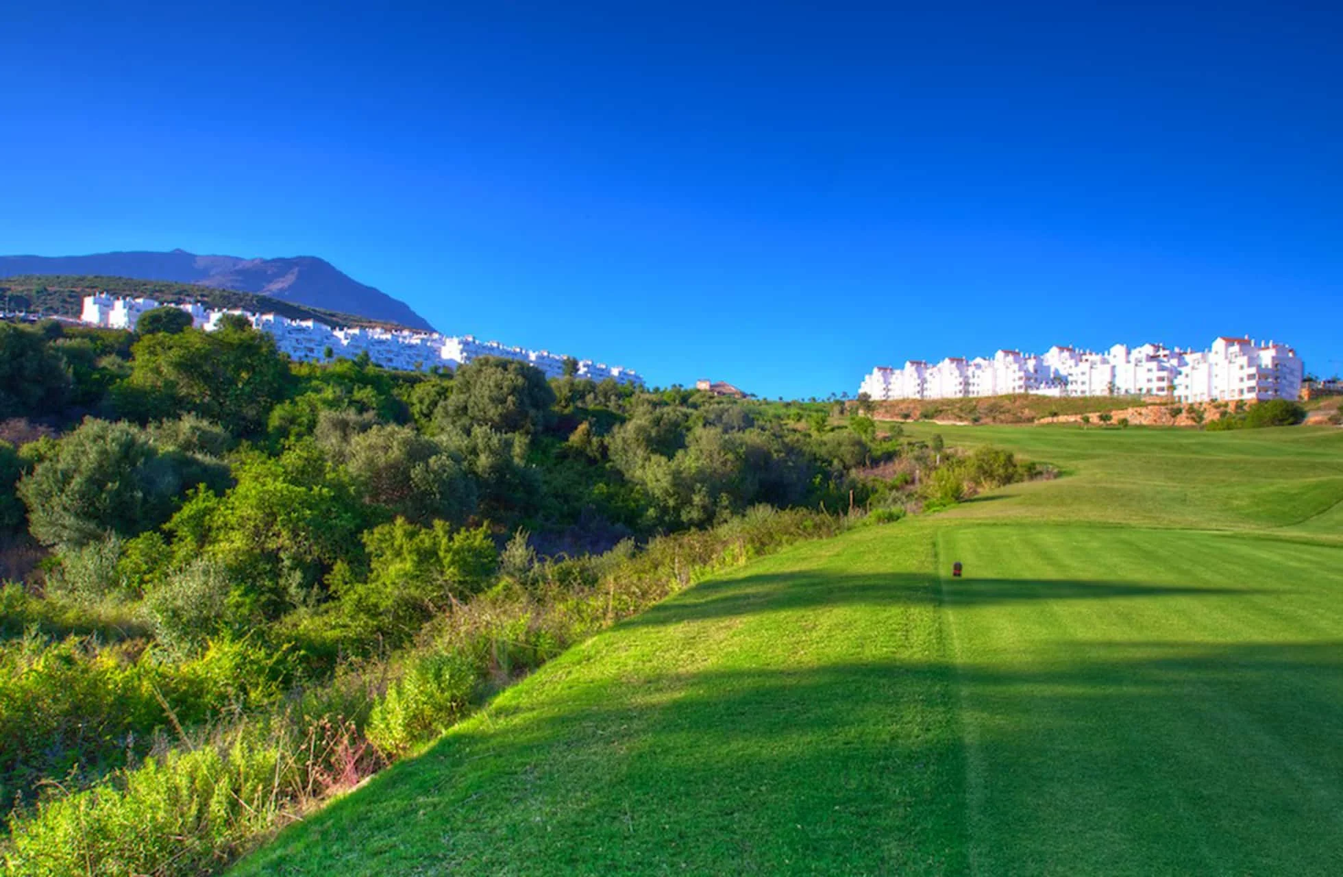 golfbane med bred fairway i kupert landskap, grøntområder langs siden og bygninger på høyder i bakgrunnen i klart dagslys