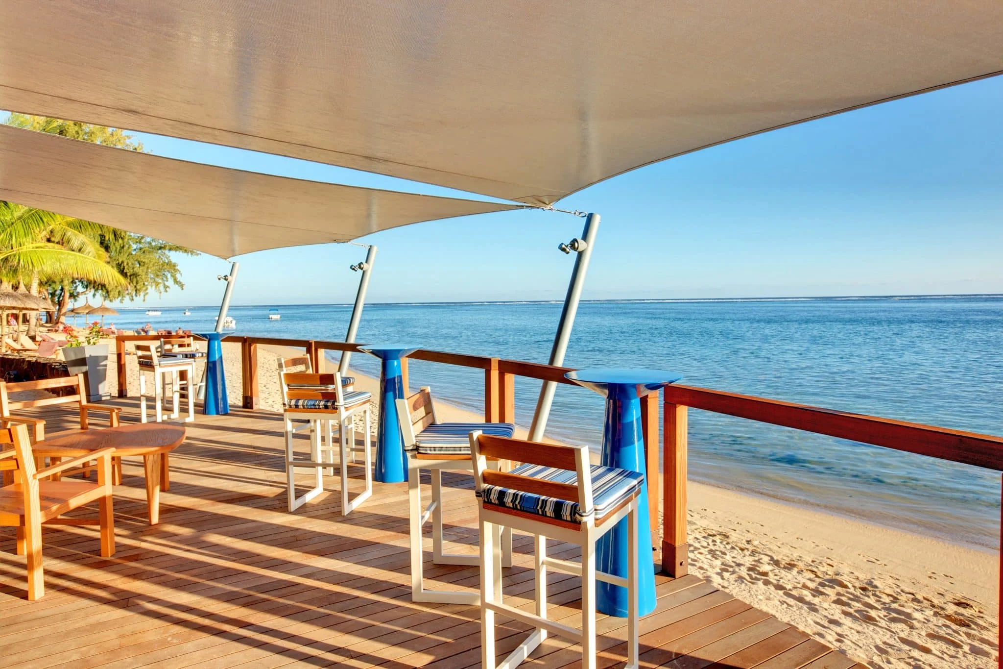 udendørs servering med træborde og stole på en terrasse ved stranden med udsigt over roligt hav og klar blå himmel.