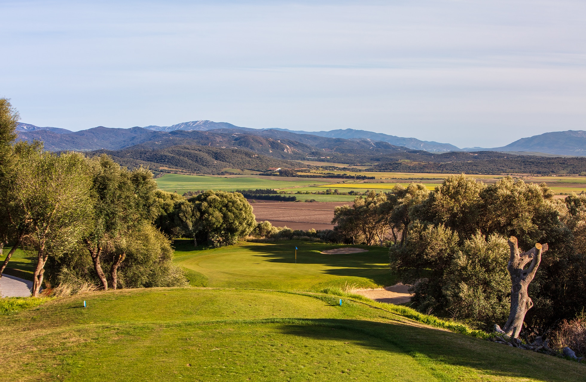golfbane med fairway i svakt bølgende landskap, omgitt av trær og med åpne jorder og fjell i bakgrunnen