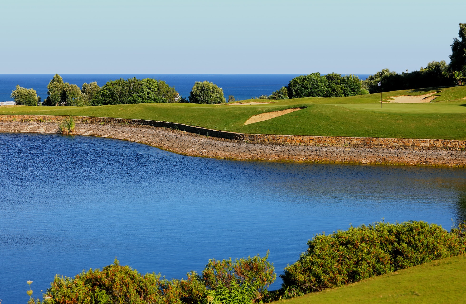 golfbane med stort vandhazard i forgrunden, green og fairway i åbent landskab med havet og horisonten i baggrunden