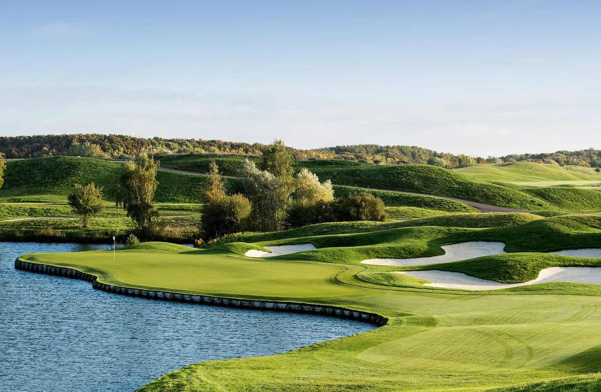 golfgreen ved vannkant med bunkere og bølgende fairwayer i åpent landskap med trær i bakgrunnen