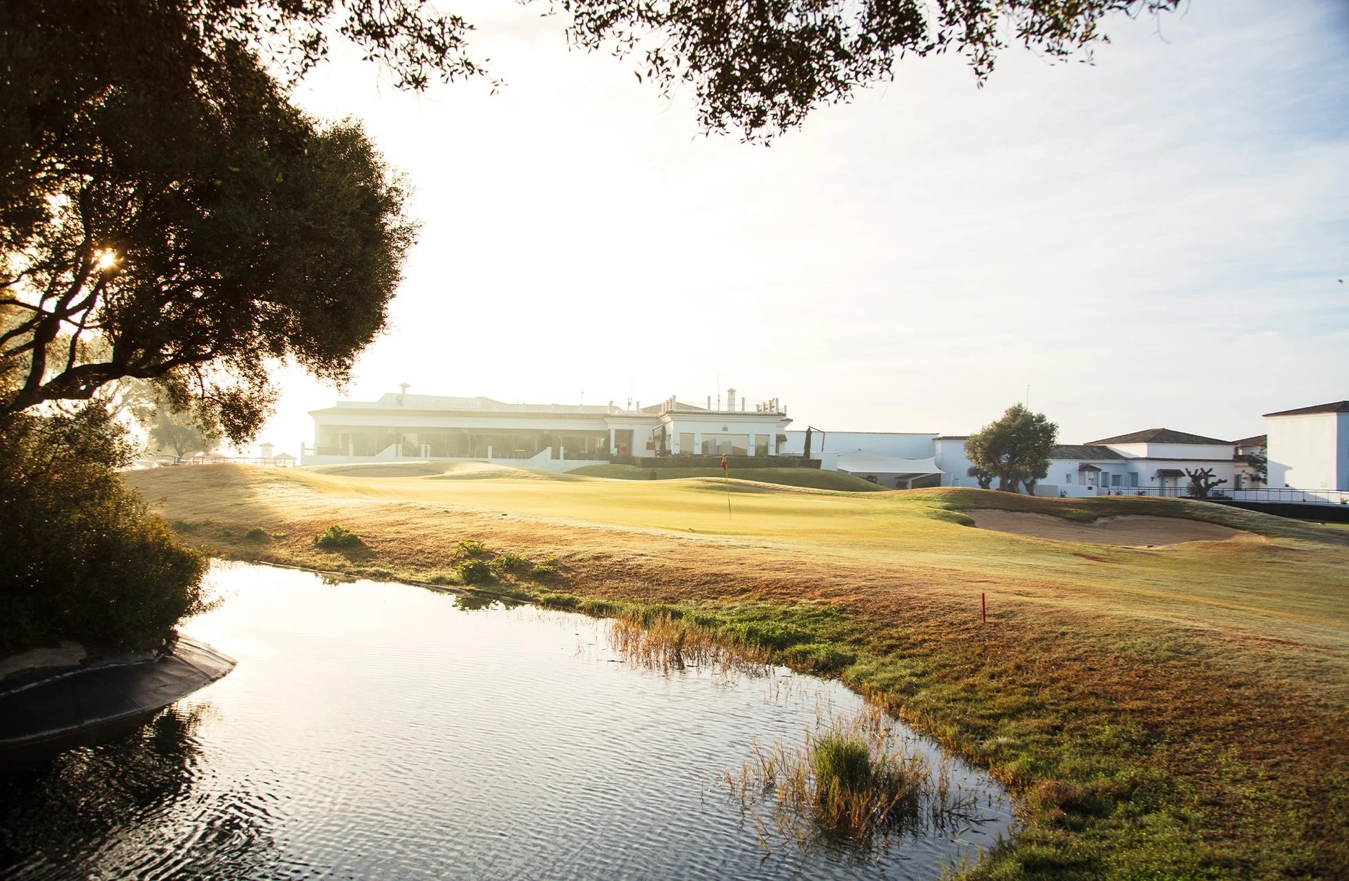 golfbane med vannhinder i forgrunnen, green og fairway i åpent landskap med bygninger og trær i varmt kveldslys
