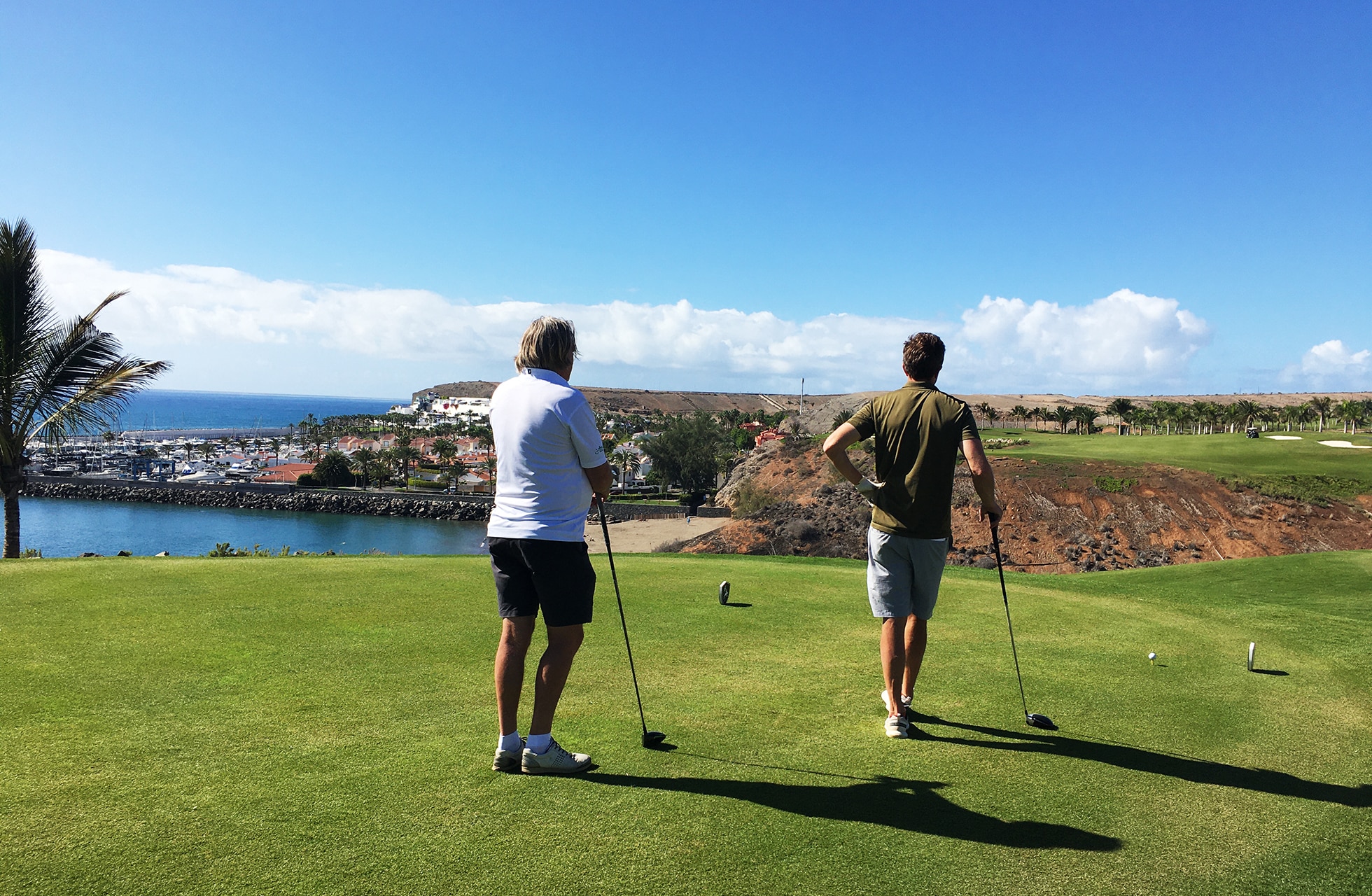 kaksi henkilöä seisoo golfin tiipaikalla valmistautumassa avaukseen, taustalla rannikko, meri ja rakennuksia