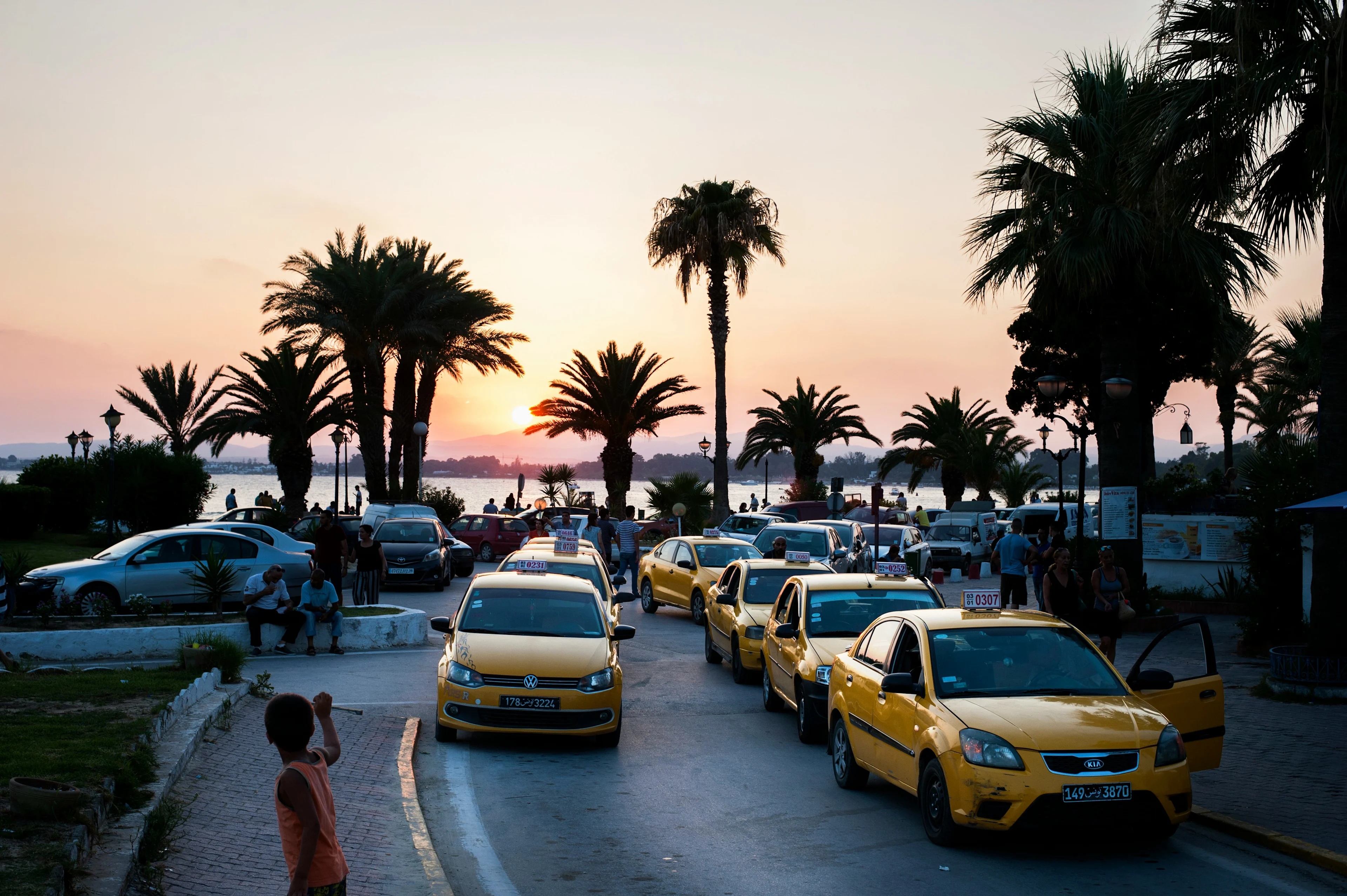 flere gule drosjer står parkert i en gate med palmer og hav i bakgrunnen under en solnedgang