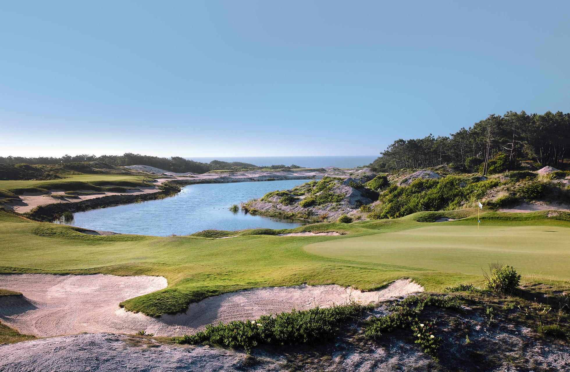 golfgreen med flagg til høyre, bunker i forgrunnen og et slynget vannhinder som går gjennom banen, omgitt av naturlig vegetasjon med havet synlig i bakgrunnen i klart dagslys