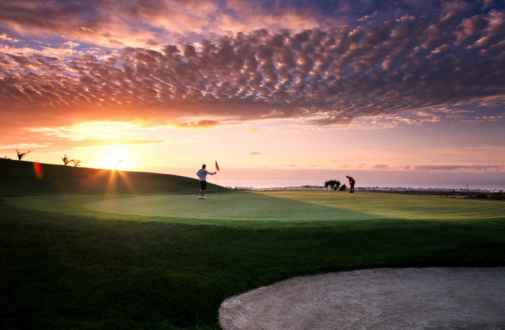 golfgreen i förgrunden med bunker, där flera golfspelare syns på banan i motljus. solen går ner vid horisonten och färgar himlen i varma toner över ett öppet landskap.