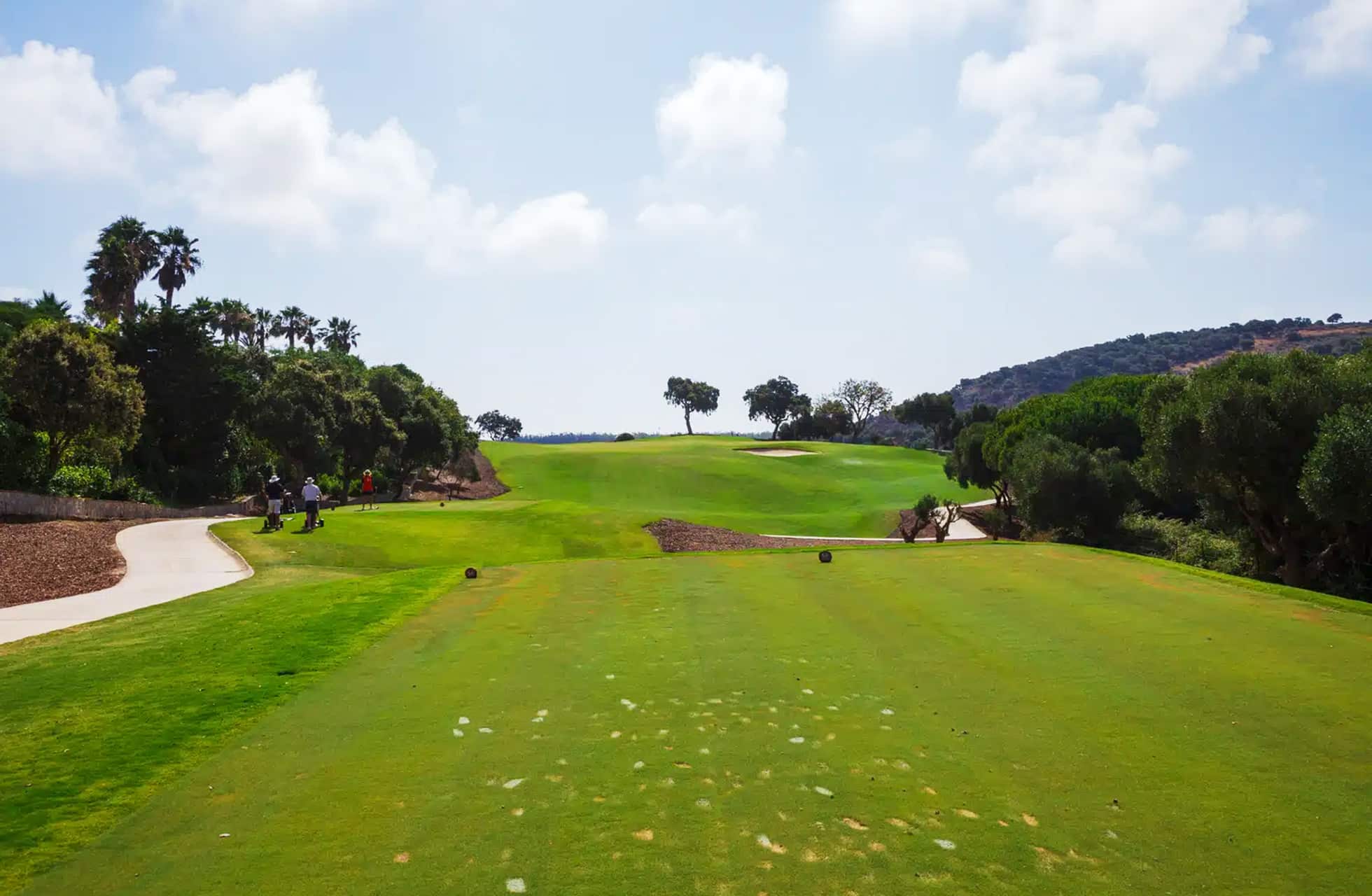 utslagssted med fairway i åpent parklandskap på san roque new course, gangvei og trær langs banen samt to golfspillere på avstand i lyst dagslys