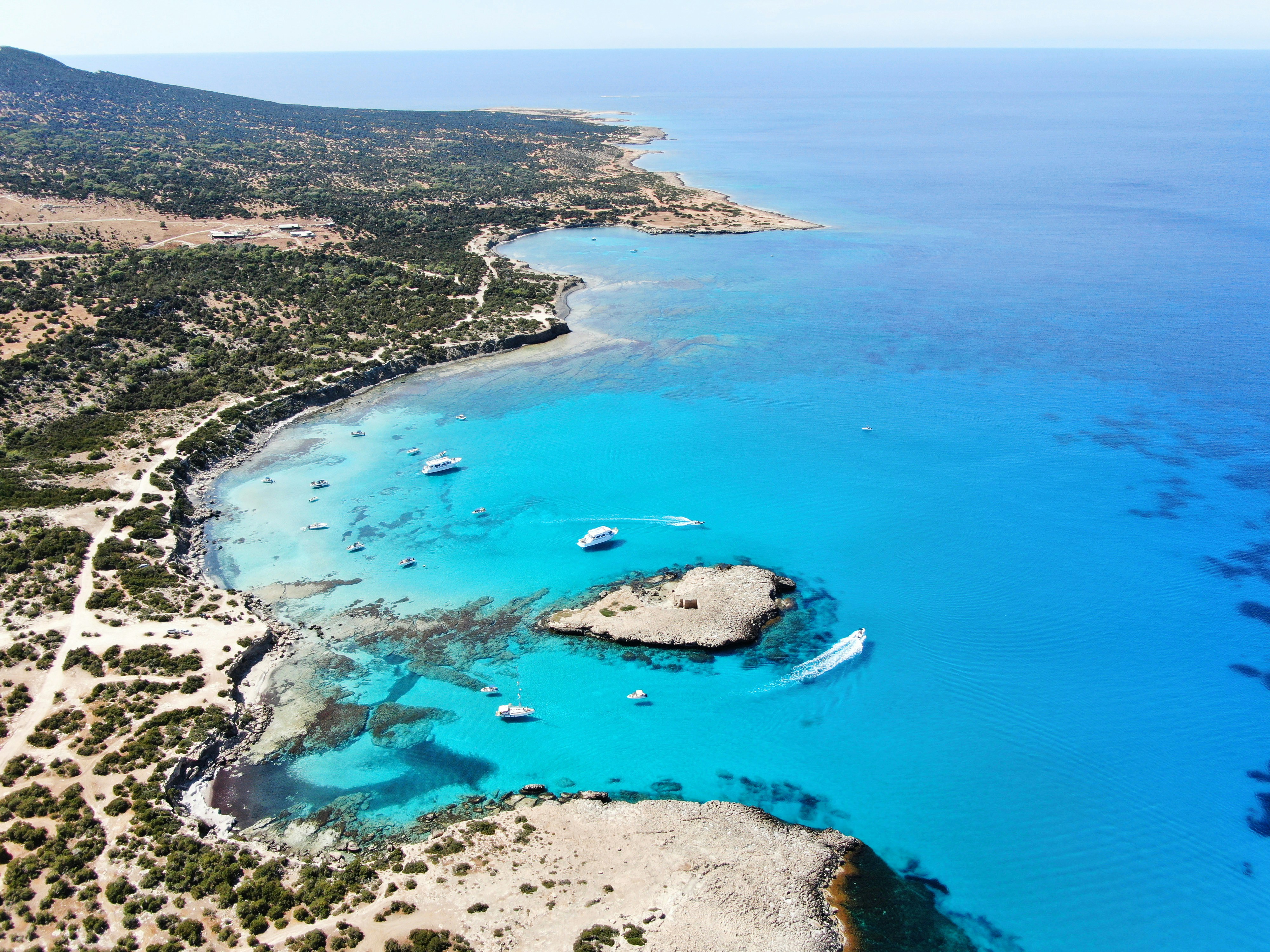 Luftfoto over en kyst med turkist hav, små øyer og båter som ligger for anker nær stranden.
