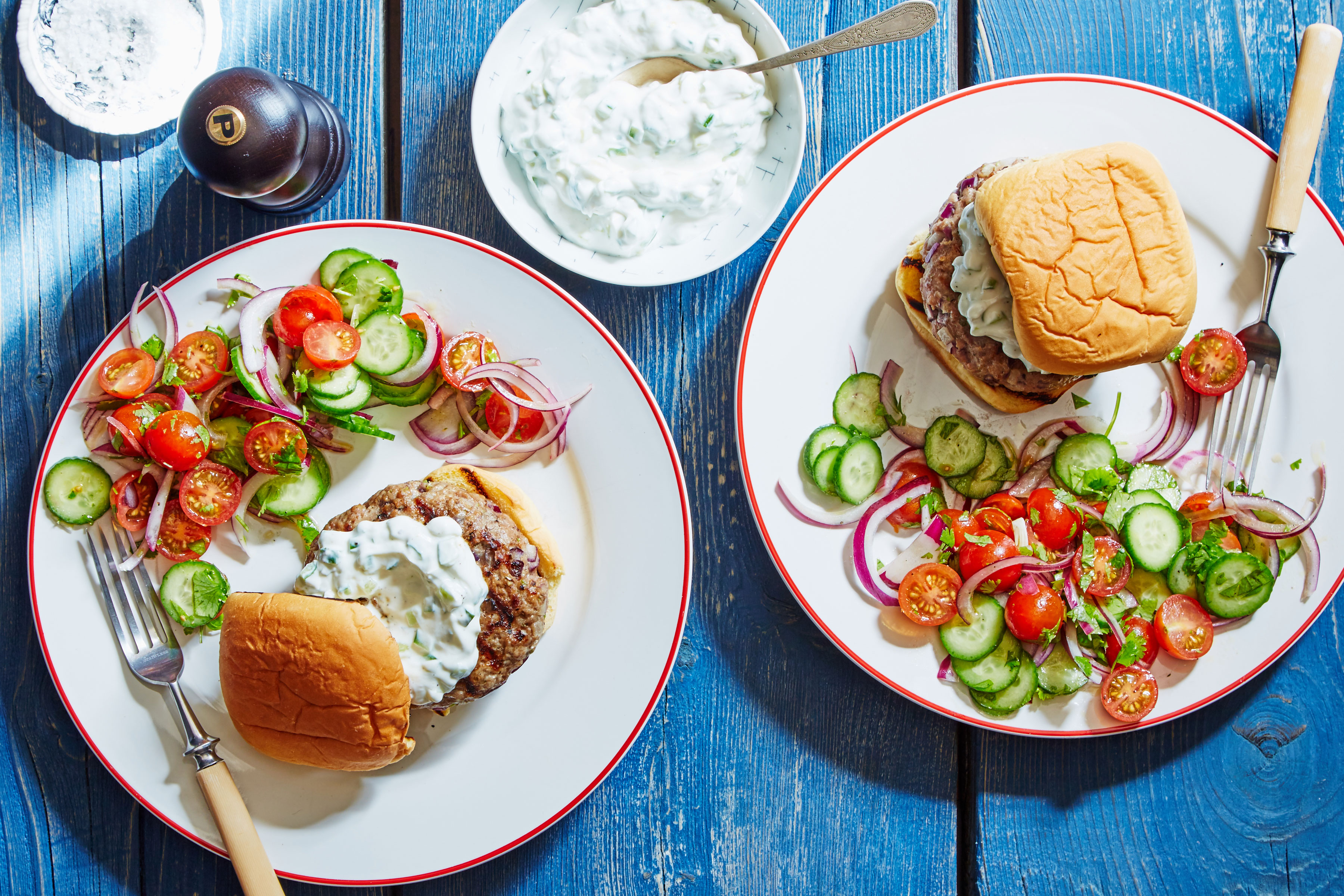 2 Pork Burgers with Cucumber Yogurt & Tomato Salad served on two white plates with a side dish of extra yogurt and a pepper grinder on a blue wood background