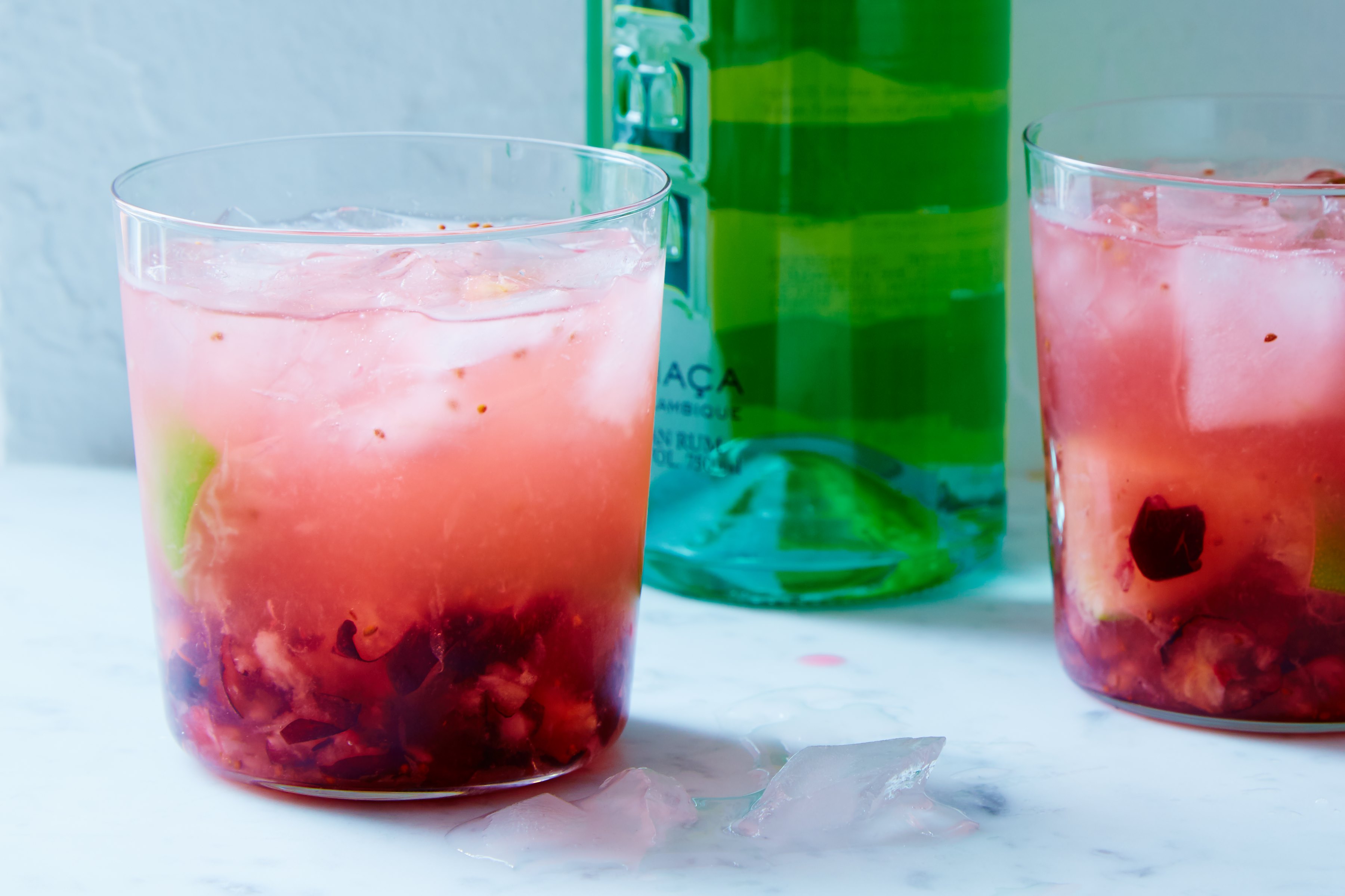 2 red cranberry caipirnha cocktails with ice in clear glasses beside a green bottle against a blue background