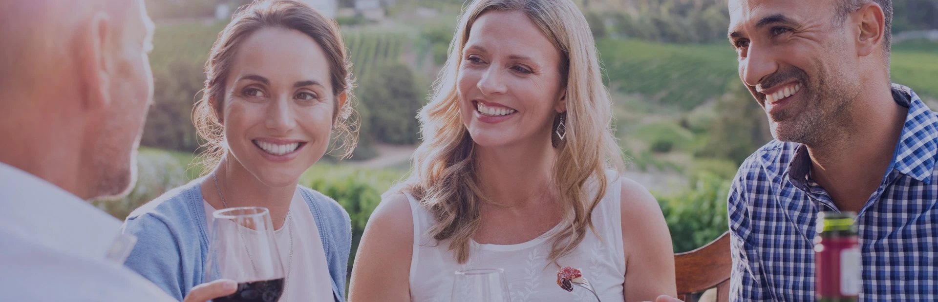 A group of friends in their forties are in a vineyard, dining al fresco, and they are all smiling, but you can't tell which one is wearing a type of partial denture.