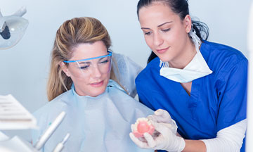 Denture Adhesive Products | Shop 4 A woman is sitting in the dentist chair with her dentist holding a set of dentures in her hands as she helps answer frequently asked questions she has about denture adhesives