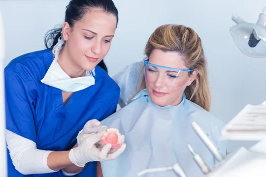 A woman is sitting in the dentist chair with her dentist holding a set of dentures in her hands as she helps answer frequently asked questions she has about denture adhesives