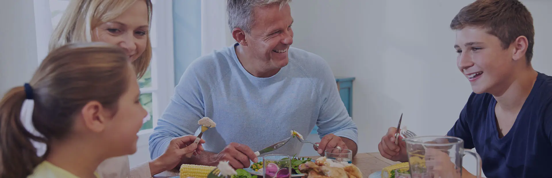 A family sit around the table eating dinner, the father has dentures but is smiling confidently and eating meat and corn on the cob, as he knows about the benefits of denture adhesive cream.  