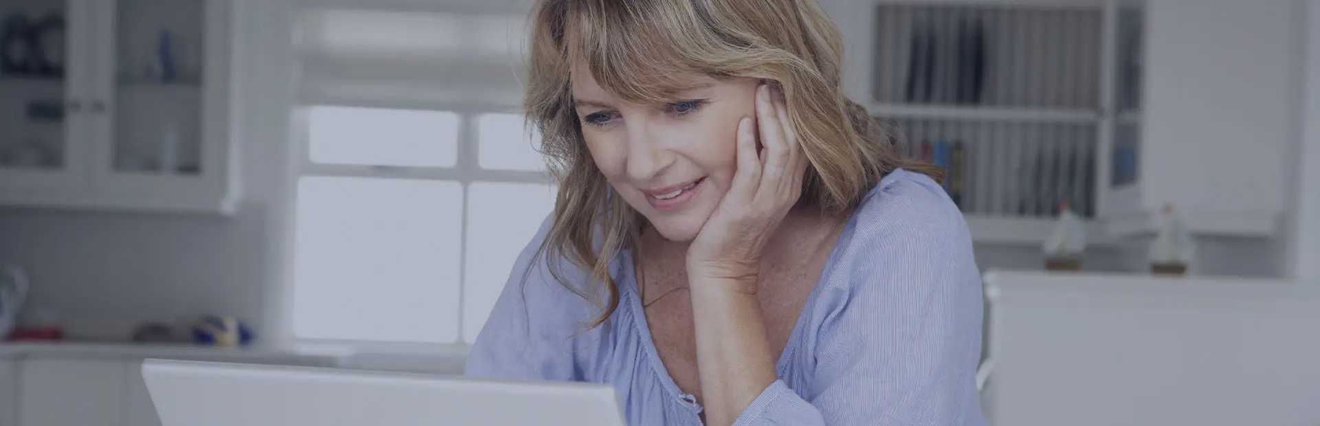 A blonde woman in her 50s wearing a blue shirt is sitting in a white kitchen researching denture stomatitis on her laptop.