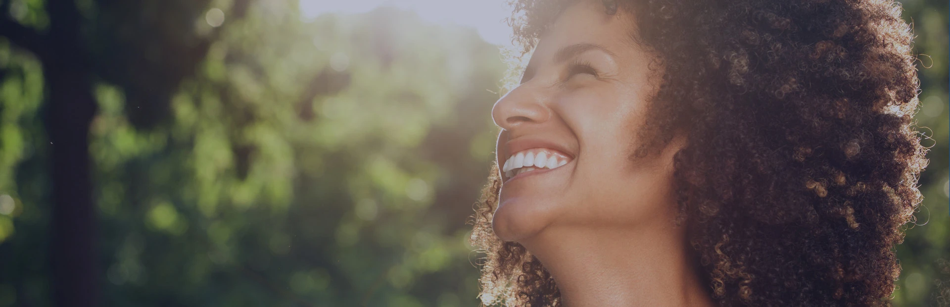 A woman in her thirties is in a sunny garden and smiling as she thinks about choosing to get dentures or dental implants. 