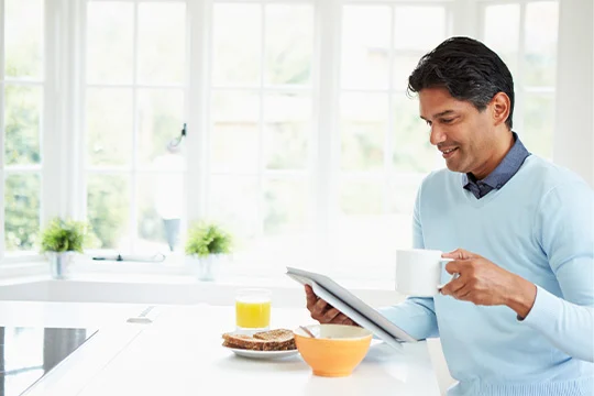 A man in his 40s is sitting in a white kitchen drinking a coffee and having breakfast while reading a brochure.