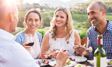 Denture Adhesive Products | Shop 6 A mixed group of friends sit in a vineyard enjoying a meal with some wine, as they know they can still have fun while living with dentures, thanks to Fixodent.