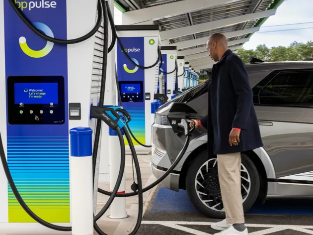 A man is connecting the charging cable plug to the port of his dark silver electric vehicle at a charging station.