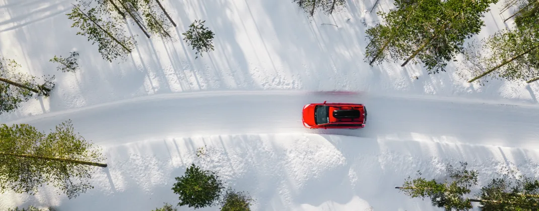 Aerial view of a red car driving along a snow-covered road surrounded by tall, snow-dusted trees in a winter landscape.