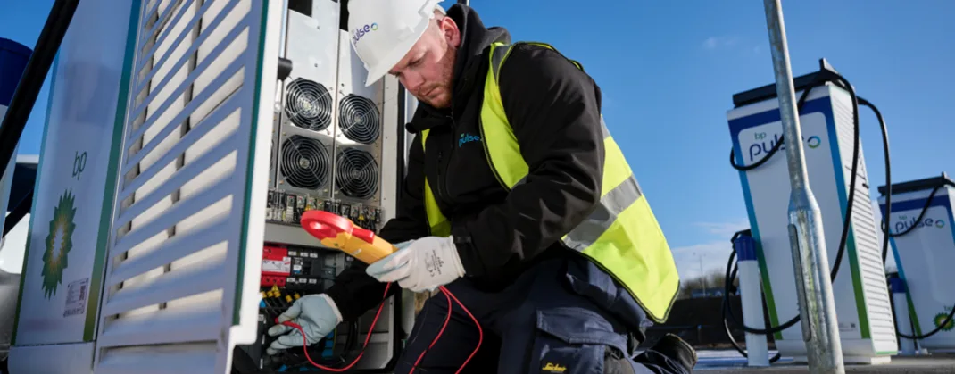 An engineer in a high-visibility vest and hard hat uses specialist equipment to run maintenance checks on a bp pulse charger. Several more bp pulse chargers are visible in the background.