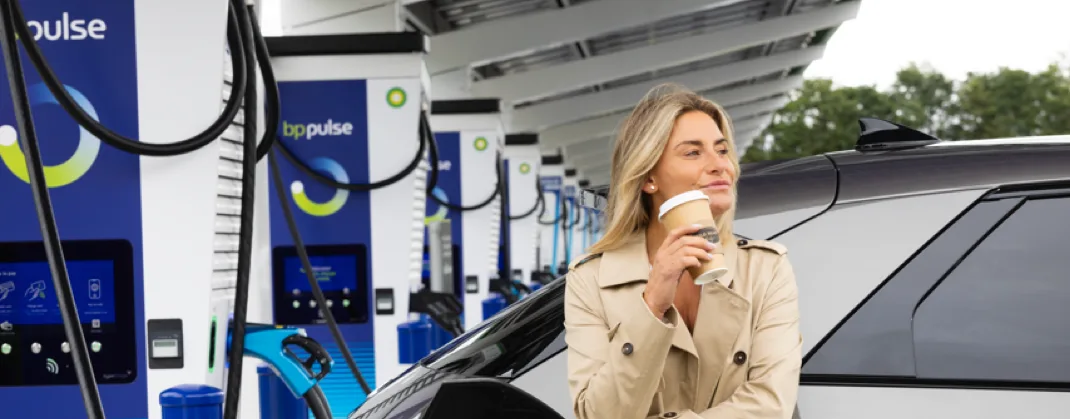A woman in coat enjoying her takeaway coffee while her EV is being charged at a bp pulse charging station. 