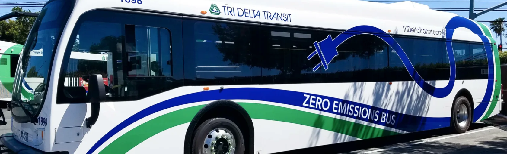A white Tri Delta Transit zero emission bus parked in a parking bay. It has green and blue connecting cables painted on it with a blue plug.