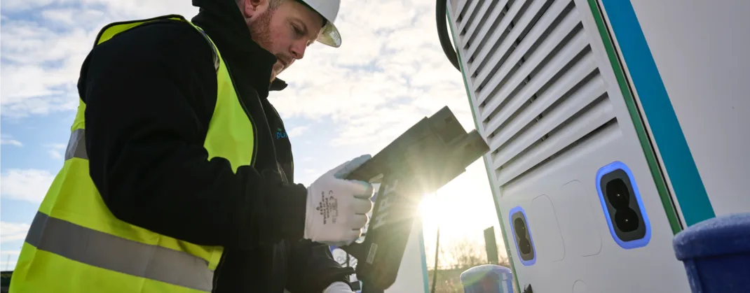 Engineer in a high-visibility vest and hard hat looking at a monitor as he runs maintenance checks on an EV charging station. 