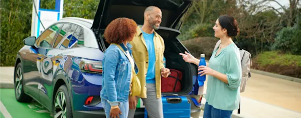 Friends standing by the open boot of a charging EV, standing and happily chatting. 