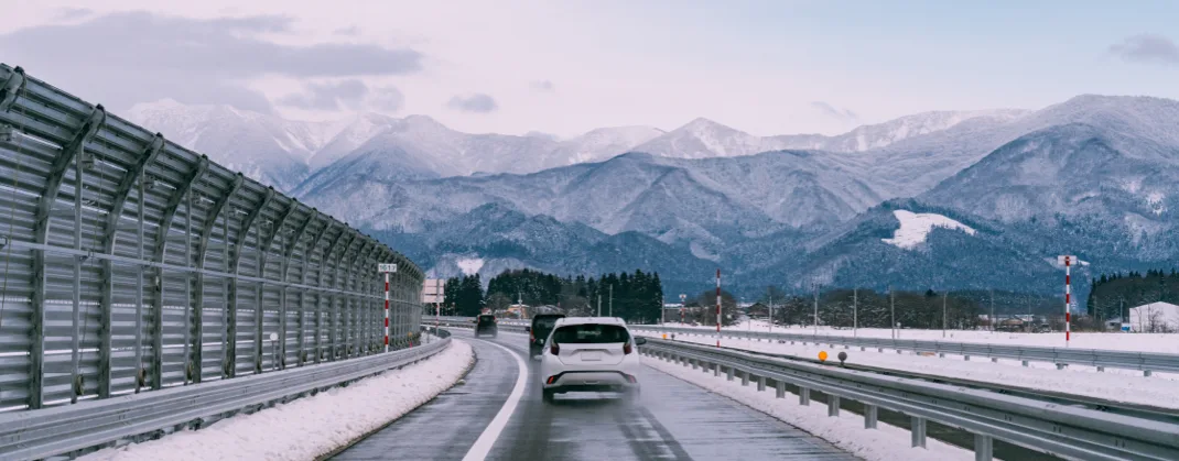 Cars driving on a road, in a winter landscape with snowy mountains in the background.