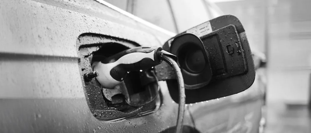 A close up of a charging cable connected to an electric vehicle, with rain drops over the car and charger cap.