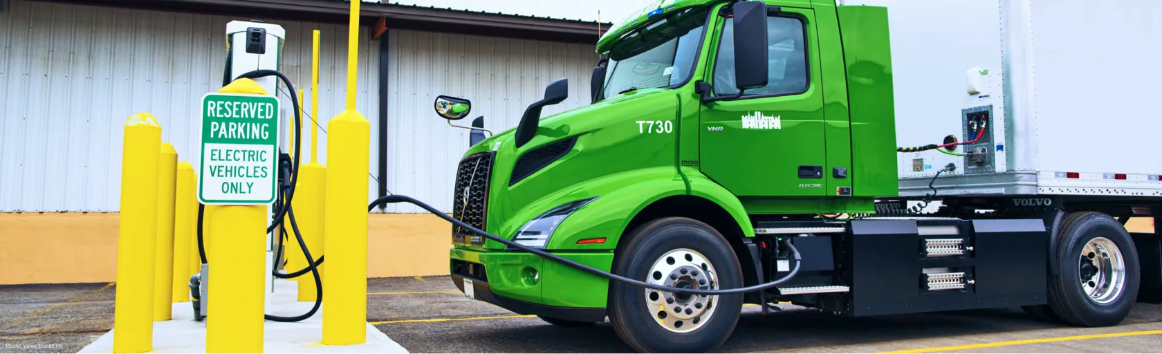 A green and white Manhattan beer truck is parked in a charging bay and plugged into an EV charger.