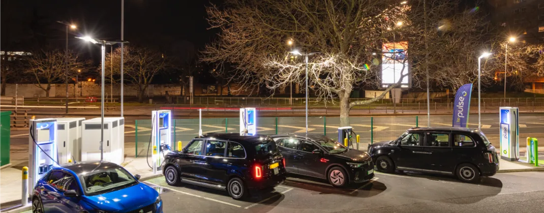 Multiple cars and taxis plugged to bp pulse chargers at a charging station at night.