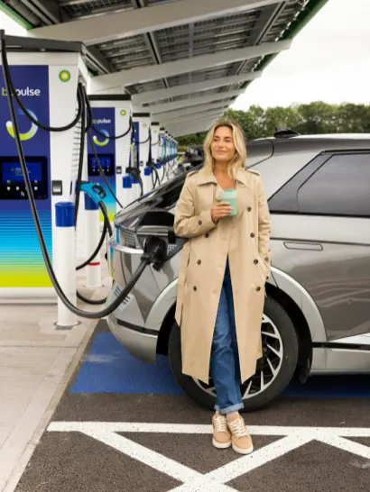 A woman holding a coffee is leaning against her silver electric vehicle while it is charging at a bp pulse charging station.