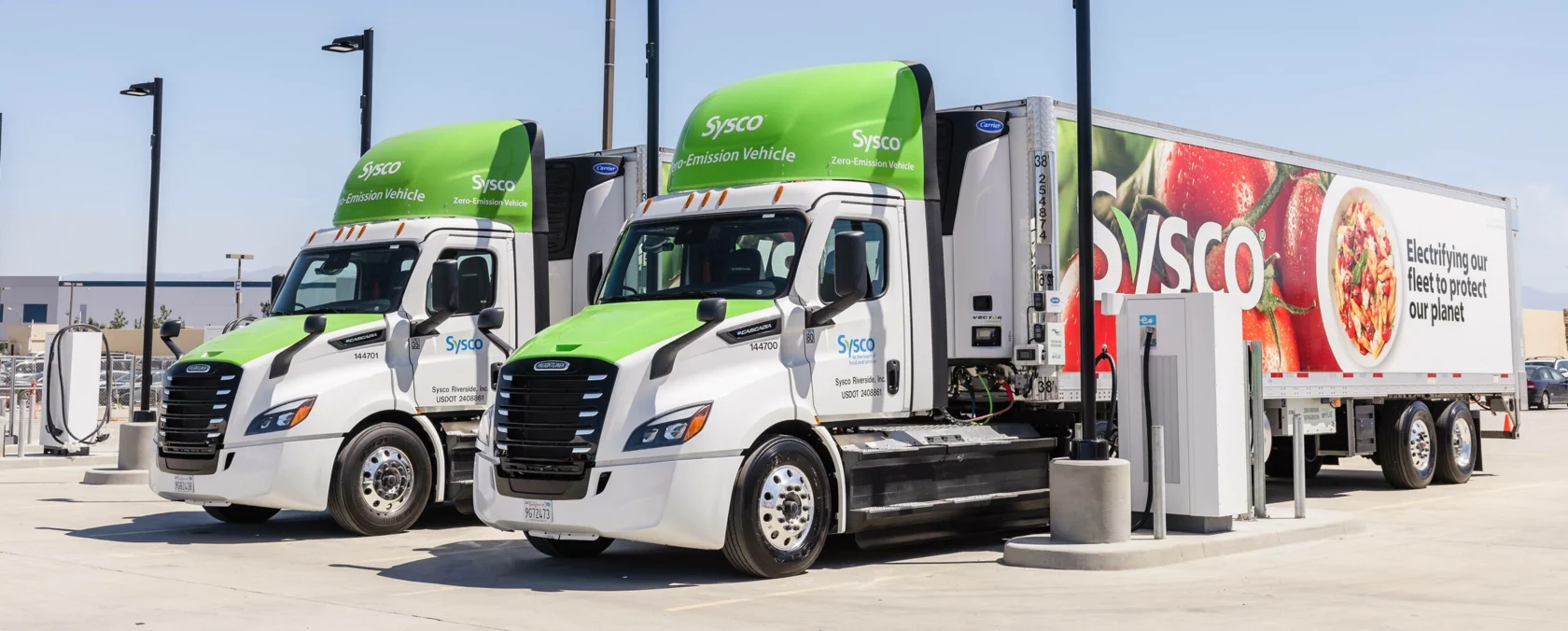 Two green and white Sysco lorries parked under some lampposts on a sunny day. 
